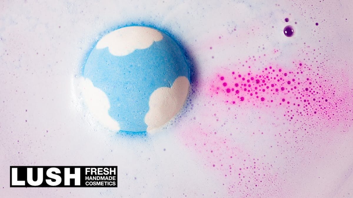 A blue bath bomb with white spots resembling clouds dissolving in foamy pink liquid. The text "Lush Fresh Cosmetics" is displayed bottom left